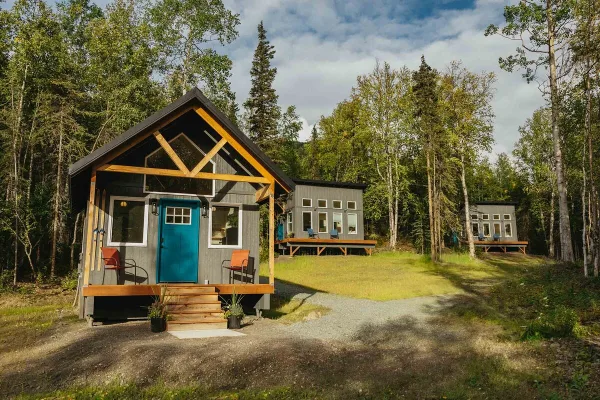 A modern wooden chalet with a blue door and exposed timber framing sits in a forest clearing, with additional guest cabins visible in the background.