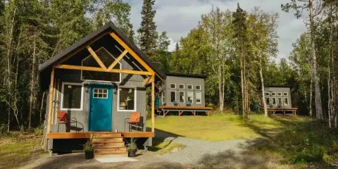 A modern wooden chalet with a blue door and exposed timber framing sits in a forest clearing, with additional guest cabins visible in the background.