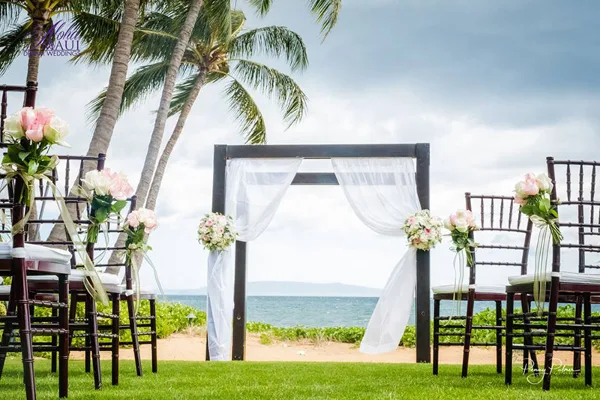 A wedding ceremony arch decorated with white drapery and flowers, set up on a grassy lawn with a view of the ocean and palm trees.