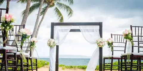 A wedding ceremony arch decorated with white drapery and flowers, set up on a grassy lawn with a view of the ocean and palm trees.