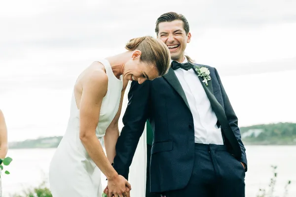 A bride and groom laughing together outdoors near a body of water.