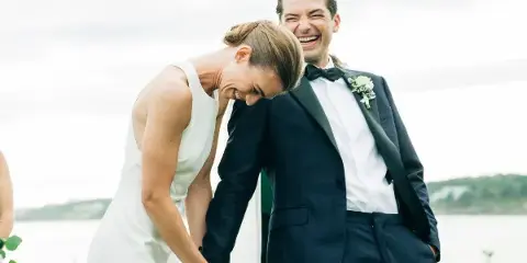 A bride and groom laughing together outdoors near a body of water.