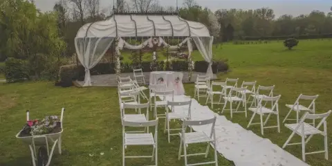 An outdoor wedding ceremony setup featuring a white canopy tent, rows of white folding chairs, and a floral aisle runner on a grassy lawn.