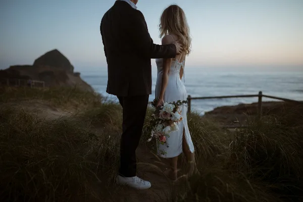 A bride and groom standing together in a coastal landscape at dusk, with the bride holding a bouquet.