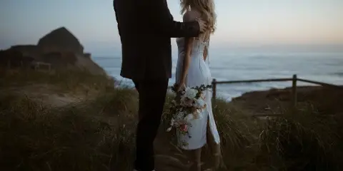A bride and groom standing together in a coastal landscape at dusk, with the bride holding a bouquet.