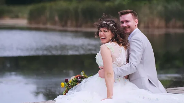 A bride and groom laughing together outdoors near a body of water, with the bride holding a bouquet.