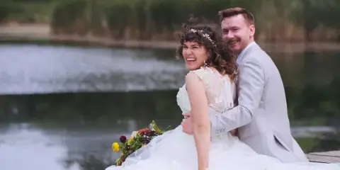 A bride and groom laughing together outdoors near a body of water, with the bride holding a bouquet.