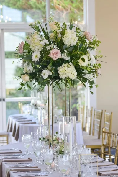 A tall, lush floral centerpiece featuring white hydrangeas and pink roses on a gold stand, set on a decorated wedding table.