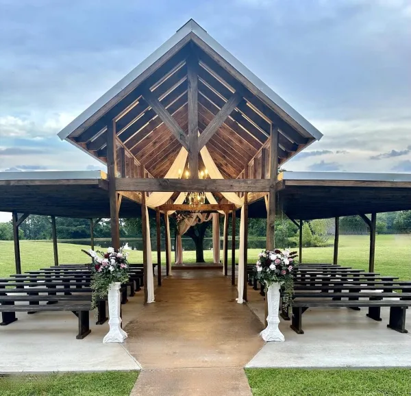 Outdoor wooden wedding pavilion with a ceremony arch, floral arrangements, and benches set against a grassy field and pond.