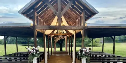 Outdoor wooden wedding pavilion with a ceremony arch, floral arrangements, and benches set against a grassy field and pond.