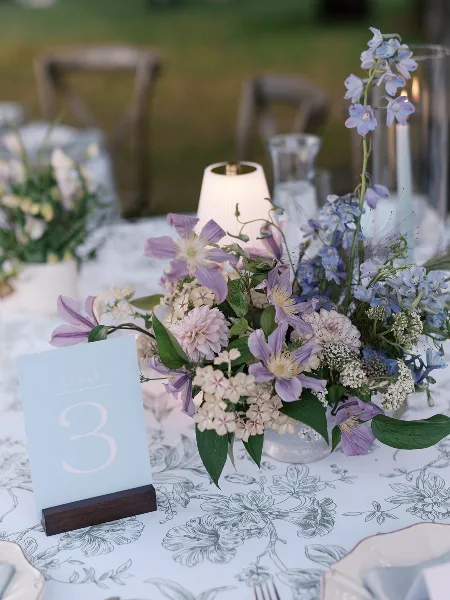 A close-up of a wedding table setting featuring a floral centerpiece with purple and white flowers, a table number card, and a patterned tablecloth.