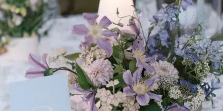 A close-up of a wedding table setting featuring a floral centerpiece with purple and white flowers, a table number card, and a patterned tablecloth.