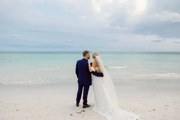 A bride and groom standing on a white sand beach facing the ocean.