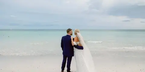 A bride and groom standing on a white sand beach facing the ocean.