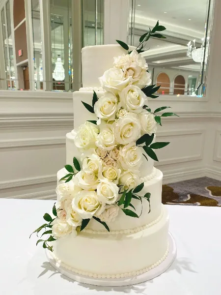 A four-tier white wedding cake decorated with cascading white roses and green leaves.