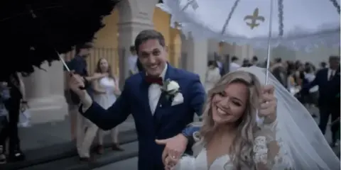 A bride and groom smiling and walking under umbrellas on a street, captured in a documentary style.