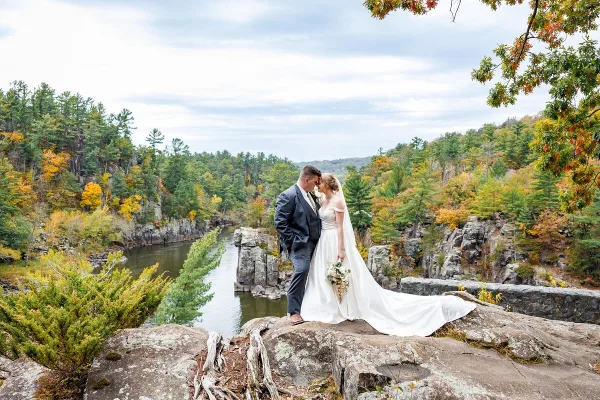 A bride and groom standing on a rocky outcrop overlooking a river and forested hills during a wedding photoshoot.