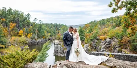 A bride and groom standing on a rocky outcrop overlooking a river and forested hills during a wedding photoshoot.