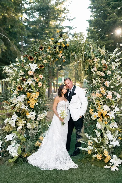 A bride and groom standing in front of a large, organic floral archway featuring white lilies, yellow flowers, and greenery.