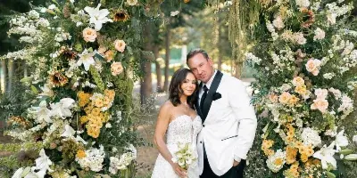A bride and groom standing in front of a large, organic floral archway featuring white lilies, yellow flowers, and greenery.