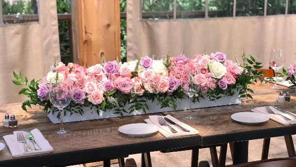 A long, low floral arrangement featuring pink and white roses and greenery in a white rectangular container, placed on a wooden table set with plates and glassware.