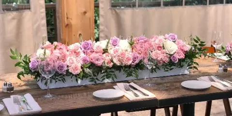 A long, low floral arrangement featuring pink and white roses and greenery in a white rectangular container, placed on a wooden table set with plates and glassware.