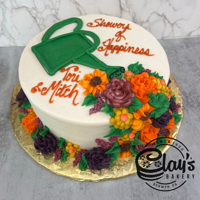 A white round cake decorated with orange and purple buttercream flowers, a green watering can, and the text 'Shower of Happiness' and 'Tori & Mitch'.