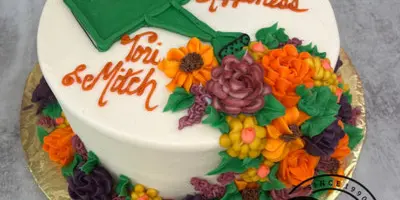 A white round cake decorated with orange and purple buttercream flowers, a green watering can, and the text 'Shower of Happiness' and 'Tori & Mitch'.