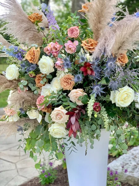 A tall, artistic floral arrangement featuring roses, pampas grass, and greenery in a white vase.
