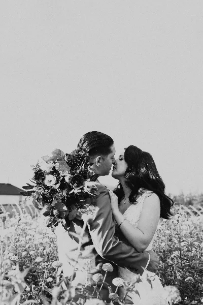 Black and white photograph of a couple in wedding attire kissing in a field of flowers.