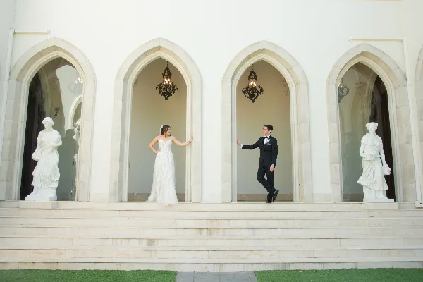 Bride in a white lace gown and groom in a black tuxedo posing in arched alcoves of a white stone venue with classical statues.
