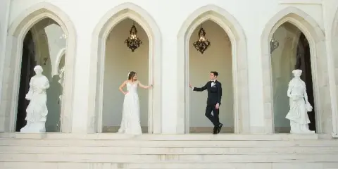 Bride in a white lace gown and groom in a black tuxedo posing in arched alcoves of a white stone venue with classical statues.