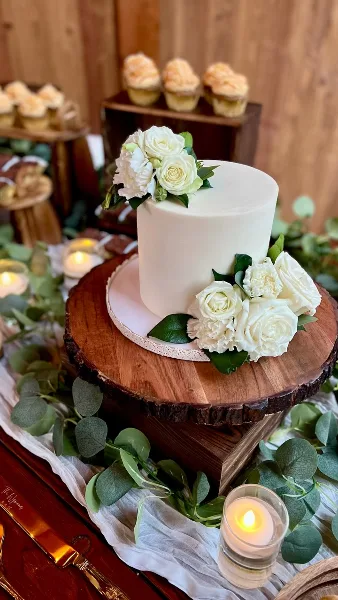 A white wedding cake decorated with fresh white roses and greenery, displayed on a rustic wooden stand with cupcakes in the background.