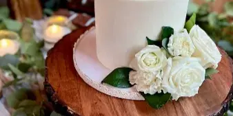 A white wedding cake decorated with fresh white roses and greenery, displayed on a rustic wooden stand with cupcakes in the background.