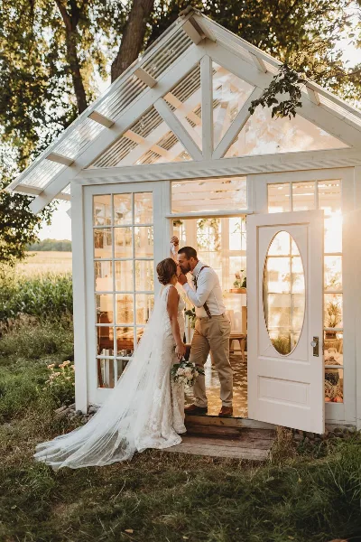 A bride and groom kissing at the entrance of a white glass-walled structure on a farm.