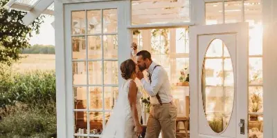 A bride and groom kissing at the entrance of a white glass-walled structure on a farm.
