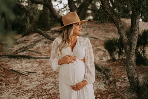 A pregnant woman in a white dress and tan hat stands outdoors in a wooded area, holding her belly in a professional portrait.