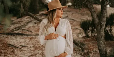 A pregnant woman in a white dress and tan hat stands outdoors in a wooded area, holding her belly in a professional portrait.
