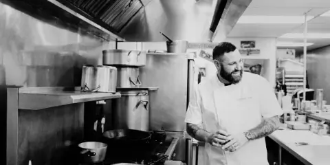 A professional chef in a white uniform standing in a commercial kitchen with stainless steel equipment.