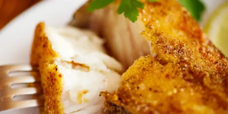 A close-up of a plated fried catfish fillet garnished with parsley and a lemon wedge.