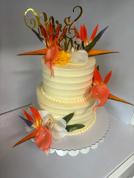 A two-tier white wedding cake decorated with tropical flowers and a gold Mr & Mrs topper.
