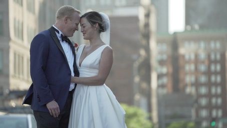 A bride and groom in wedding attire standing close together outdoors with a city background.