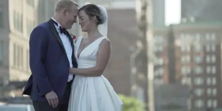 A bride and groom in wedding attire standing close together outdoors with a city background.