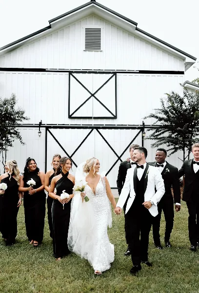 A wedding party walking on a lawn in front of a large white barn with black trim.