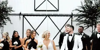 A wedding party walking on a lawn in front of a large white barn with black trim.