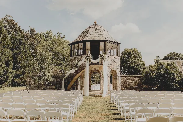 Outdoor wedding ceremony setup with rows of white chairs facing a stone tower gazebo decorated with white drapery.