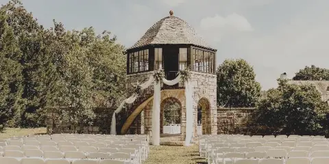 Outdoor wedding ceremony setup with rows of white chairs facing a stone tower gazebo decorated with white drapery.