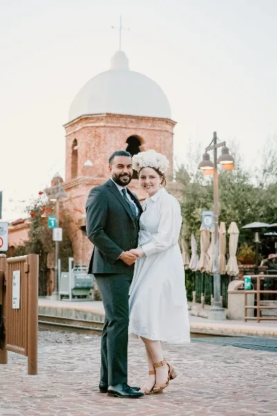 A wedding couple posing outdoors in front of a historic brick mission-style building with a white dome.