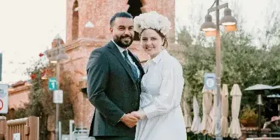 A wedding couple posing outdoors in front of a historic brick mission-style building with a white dome.