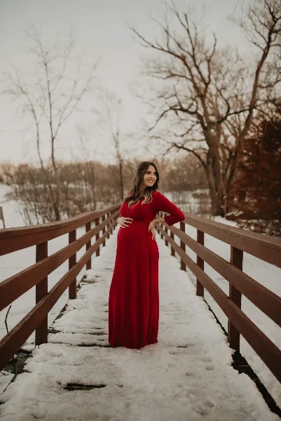 A woman with styled hair and makeup wearing a long red dress standing on a wooden bridge in a snowy landscape.